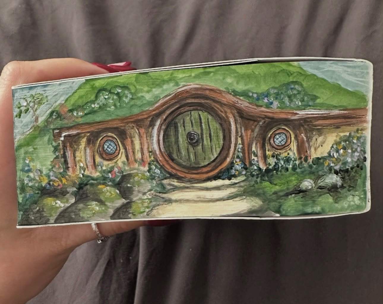 Fore-edge painting on a paperback of Bag End’s round green door in a grassy hill, with round windows and a path through the flowers.