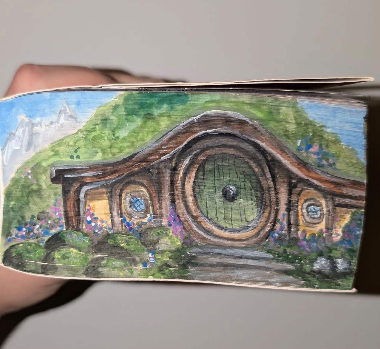 Fore-edge painting of Bag End in the Shire, its round door set into a green hill with purple and blue flowers and the Misty Mountains beyond.